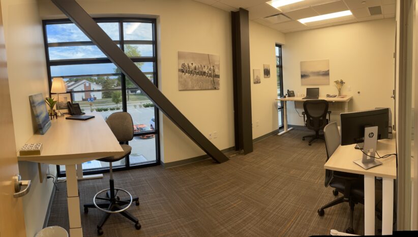 Contemporary office workspace with desks, computers, and large windows providing natural light.