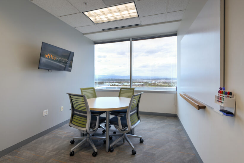 Modern office meeting room with round table and green chairs, large window with city view, Office Evolution branding on wall mounted TV.