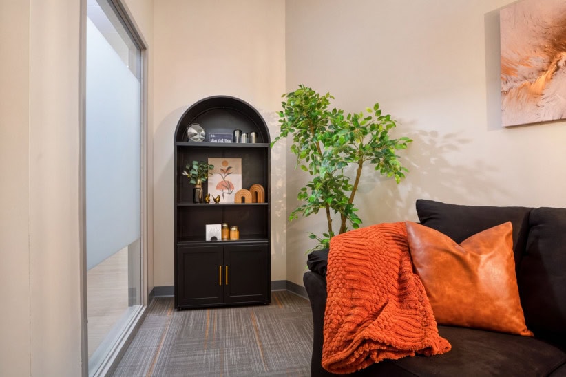 Modern office lounge with black sofa, orange pillows, and a green plant.