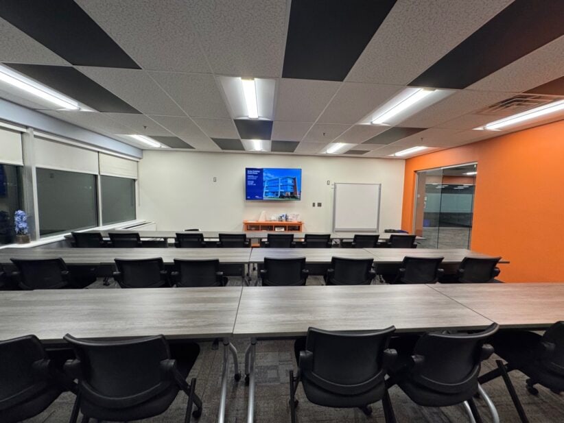 Photograph of a training room with orange wall, window views, and checkerboard ceiling tiles. seating for 20 or more individuals.