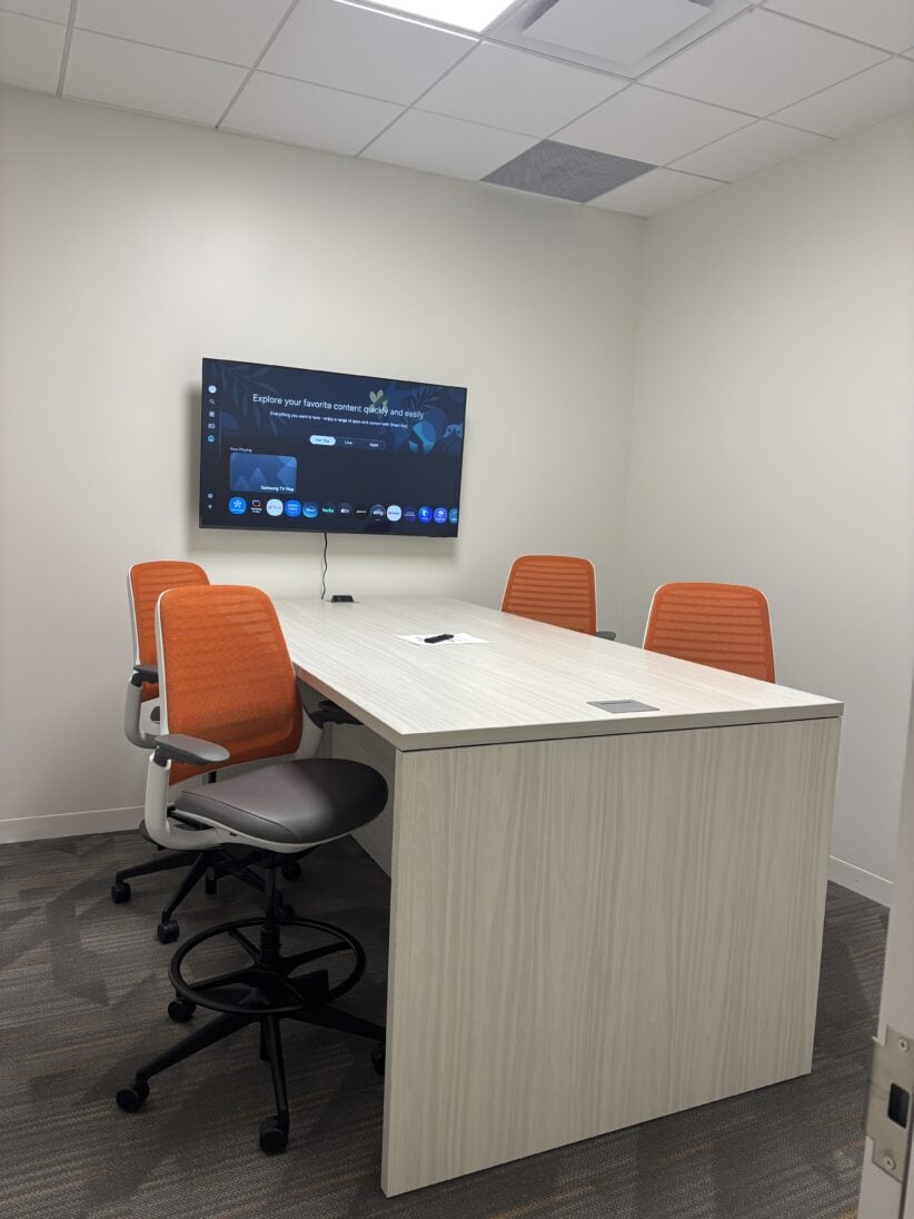 Modern office meeting room with conference table, orange chairs, and wall-mounted TV for collaborative workspace.
