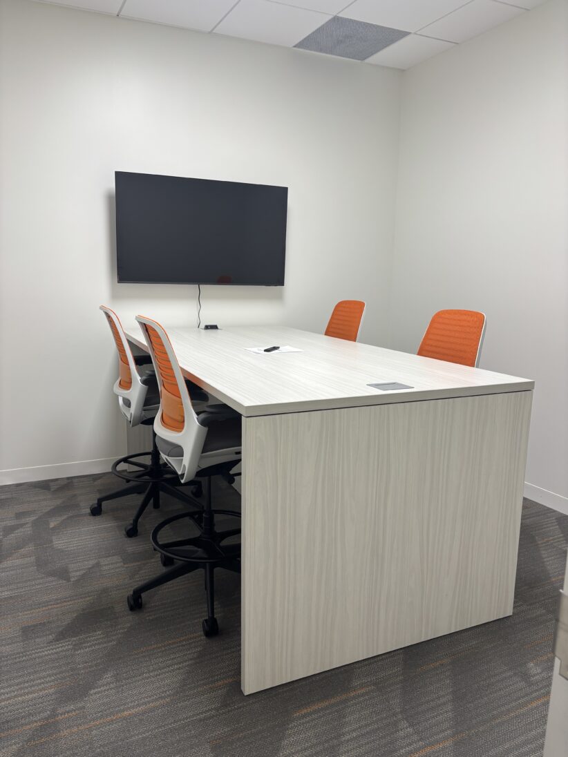 Modern collaborative office space with white table, orange chairs, and wall-mounted TV for meetings and remote work.