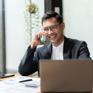 image of man with glasses sitting at office desk with is laptop, and he is talking on the phone