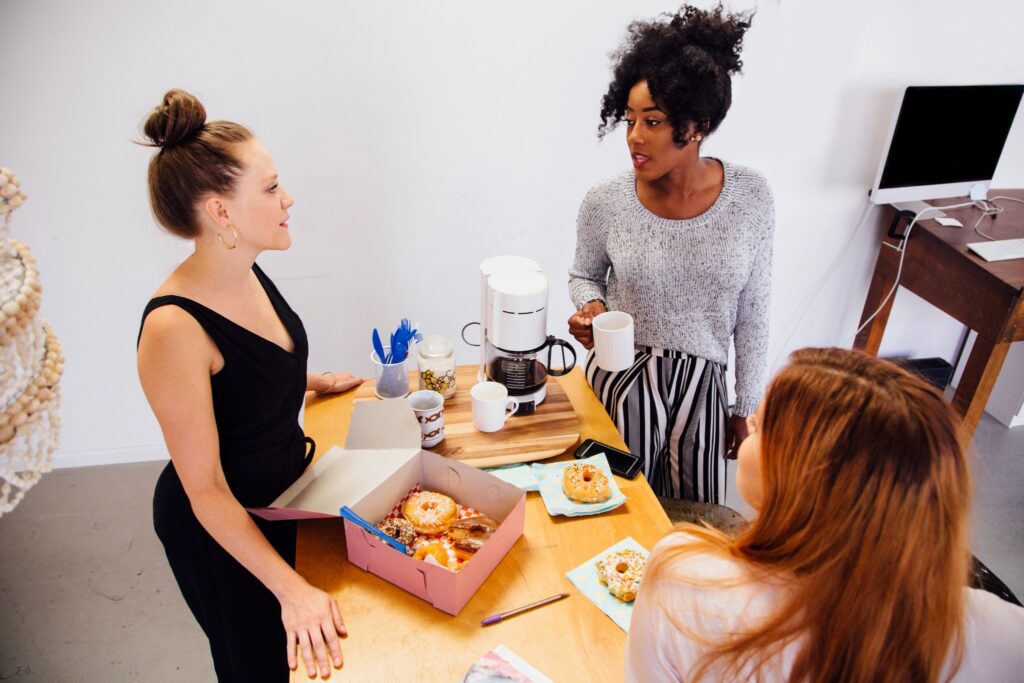 Coworking space etiquette requires communication and attentiveness. Image of three women eating donuts in shared office kitchen space.