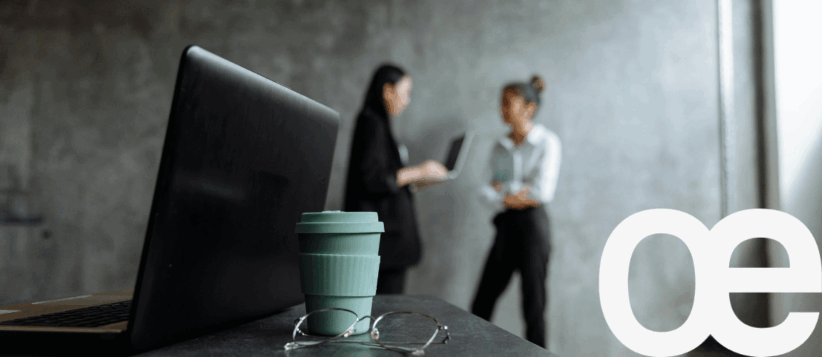 Coffee cup and glasses on office desk with laptops and two women chatting in background, modern coworking space, Office Evolution.