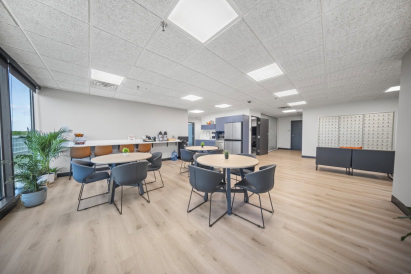 Modern office lounge with tables, chairs, and a coffee station in a bright space.