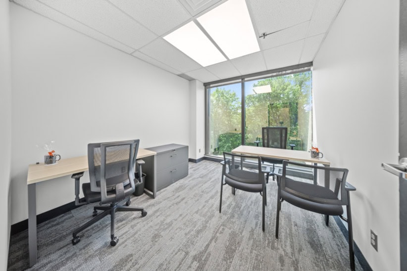 Modern office space with desks, chairs, and large window overlooking greenery.