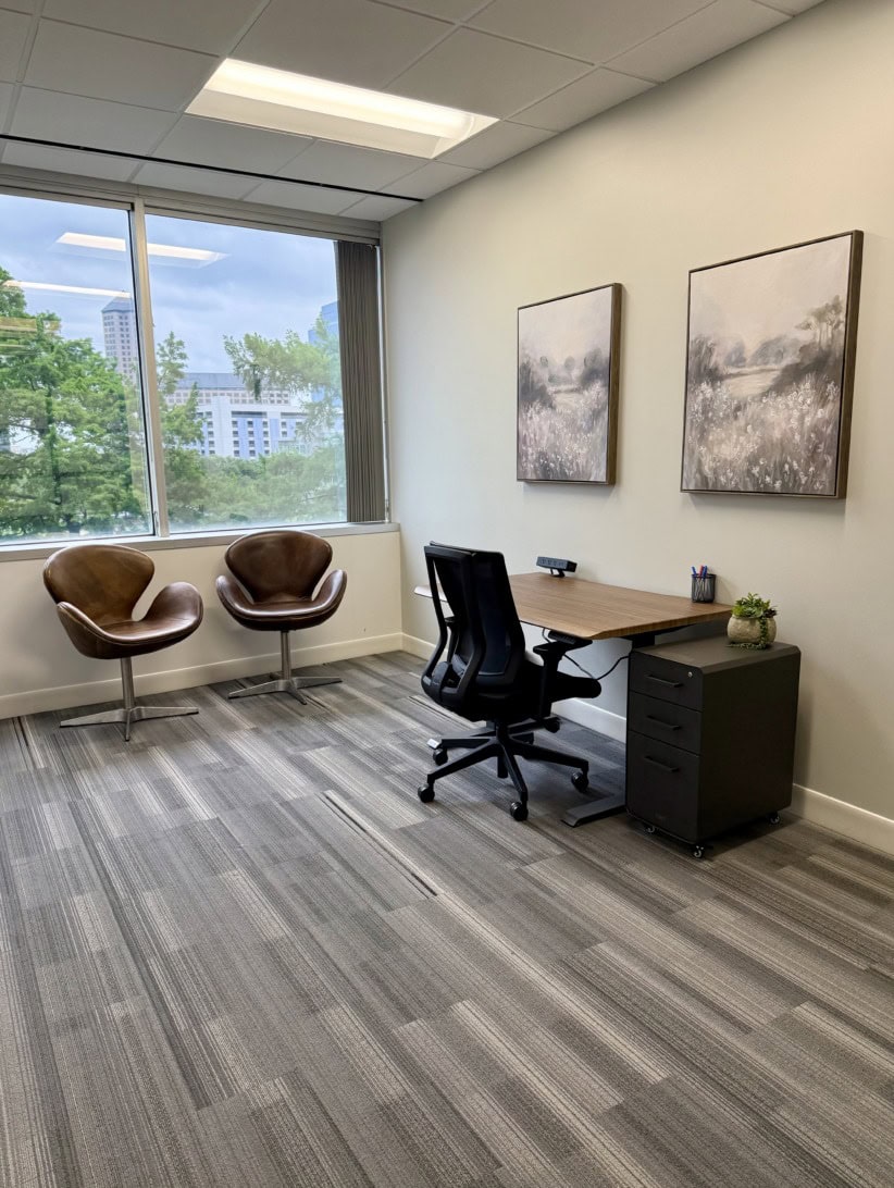 Modern office space with a desk, chair, and two armchairs near a large window.