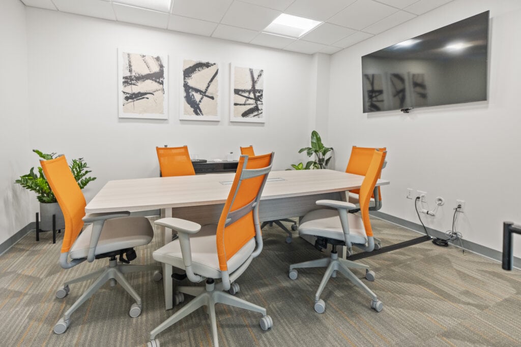 Modern office meeting room with orange chairs and a large TV screen.