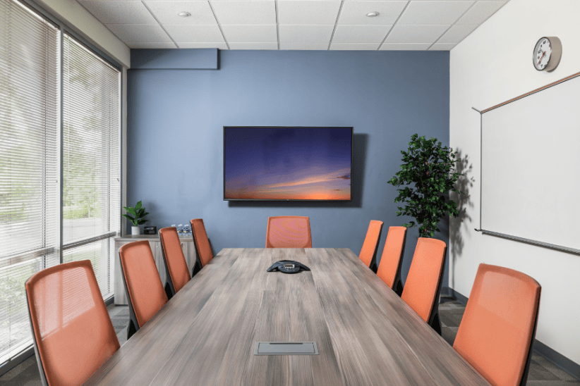 Modern conference room with a large table and orange chairs, TV on blue wall, and large windows.