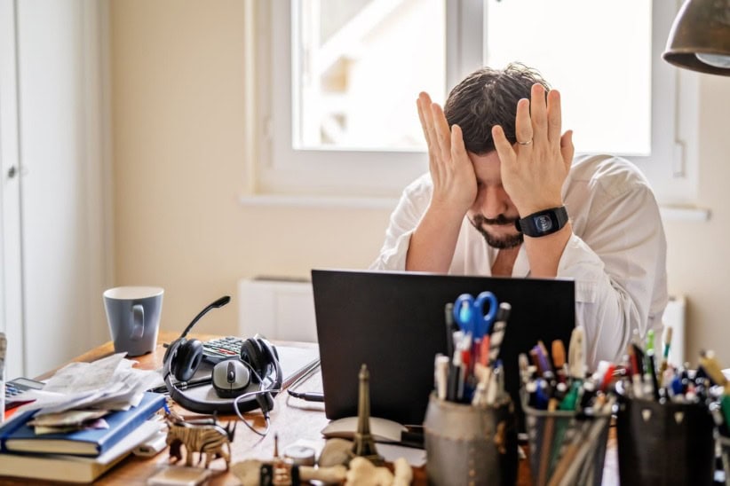 Frustrated man working from home with head in hands at cluttered desk.